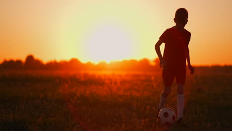 Boy-football-player-at-sunset-runs-with-a-soccer-ball-on-the-field-training-dribbling-from-dawn-to-dusk.-Way-to-success.-The-concept-of-achieving-the-goal-of-a-successful-athlete